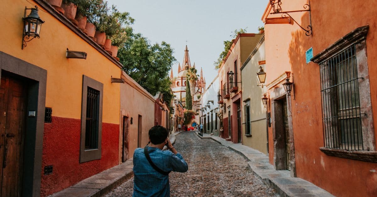 A man photographs a cobbled street of orange and red houses in San Miguel de Allende, Mexico, with a church spire beyond.