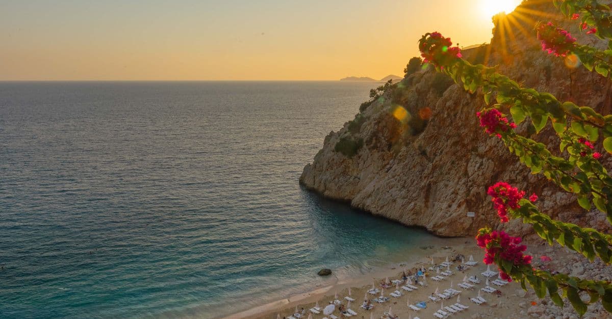 Sunset over Kaputaş Beach in Antalya, Turkey, with pink flowers above a cove lined with sun loungers.